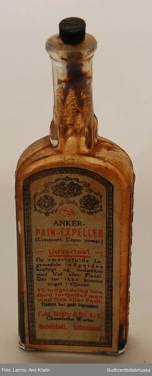 Medisinflaske, rektangulær og flat i formen. På mørkbrun trekork er avtegnet et anker, det samme på baksiden av flasken. 