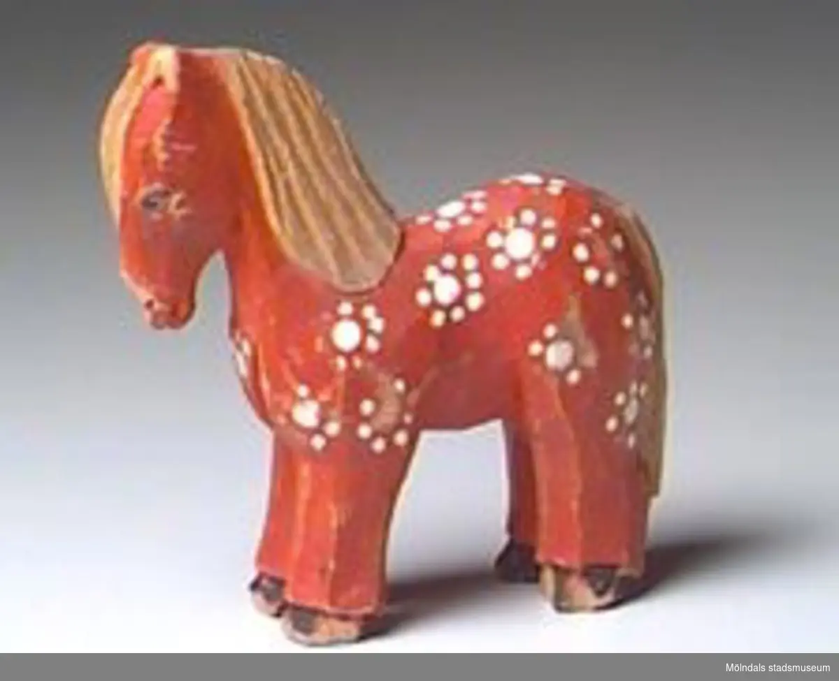 Rödmålad häst med vita prickar, svarta hovar och ofärgad svans och man. En bit av mulen är borta.Förvaras i en brun kartong.