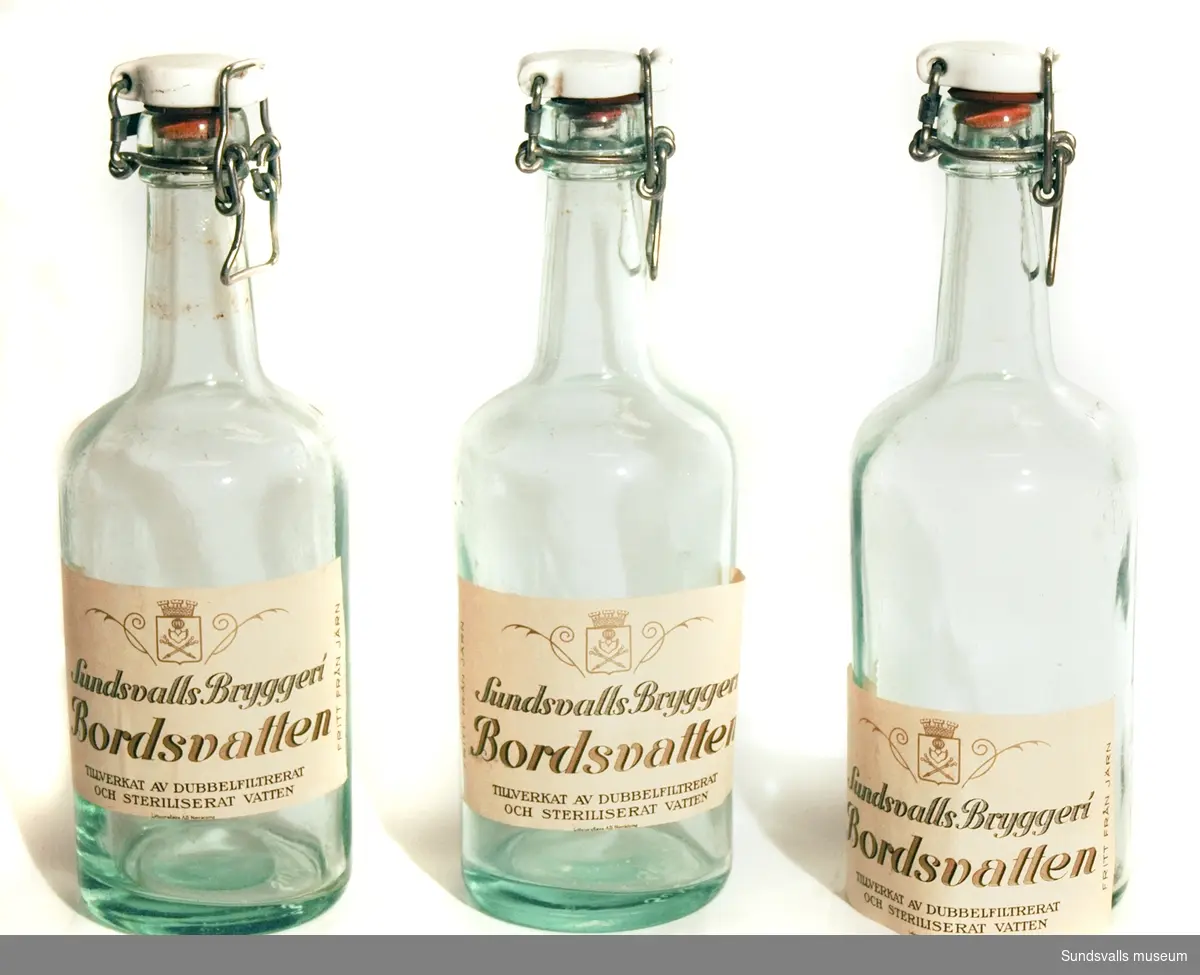 Tre flaskor med patentkork för bordsvatten från Sundsvalls Bryggeri. Flaskorna är gjorda i ofärgat glas och är försedda med vita etiketter med texten 'Sundsvalls Bryggeri, Bordsvatten, TILLVERKAT AV DUBBELFILTRERAT OCH STERILISERAT VATTEN, FRITT FRÅN JÄRN'. Etiketterna är tillverkade hos Lithografiska AB i Norrköping.