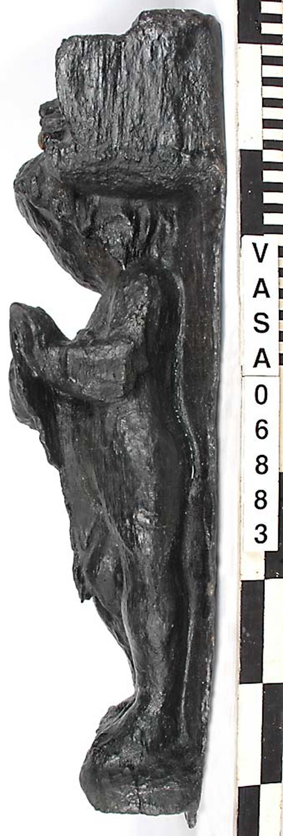 Skulptur föreställande en barnfigur spelande en harpa.
Instrumentet hålls med bägge händerna. Huvudet är vridet något åt höger. Kroppstyngden vilar på vänster ben medan höger ben är böjt. Vid höger sida syns en mantel. Ovanför hjässan sitter en snedställd äggstavslist. Figuren står på ett minimalt underlag som troligen avser att föreställa ett stycke naturlig mark. Skulpturens baksida är slät.
Skulpturen är mycket sliten.

Text in English: This is a sculpture of a child playing a harp.
The instrument is held in both hands. The head is turned slightly to the right. The weight of the body rests on the left leg, the right leg is bent. A mantle is to be seen to the figures right side. Just above the head the back board comes foreward at right angles and the front of the board is wide and is decorated with an egg moulding design which is placed diagonally across it with the right-hand side lower than the left-hand side, when the sculpture is viewed from the front. The figure stands upon a small flat plinth. The back of the sculpture is smooth.
The sculpture is very  worn.