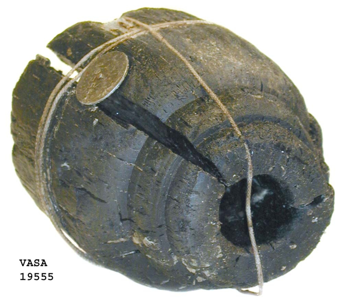 Kraftigt eroderad kolv till ansättare. Kolven består av ett cylindriskt svarvat trästycke med ett centrumhål för skaftet. I kolvens övre ände, mot skaftet till, är kolven profilsvarvad. Föremålet är brutet i två delar vilka hålls samman med snöre.
