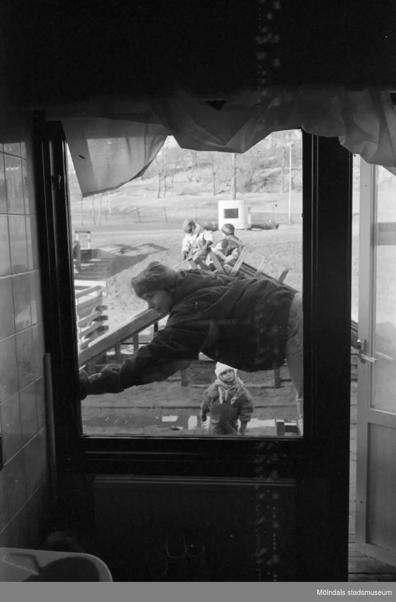 Bild fotograferad inifrån och ut genom ett fönster från Katrinebergs daghem. En balkongdörr till höger är lite öppen. Utanför syns en vuxen person med mössa som lutar sig över ett staket. Lite längre bort ses vinterklädda, lekande barn.