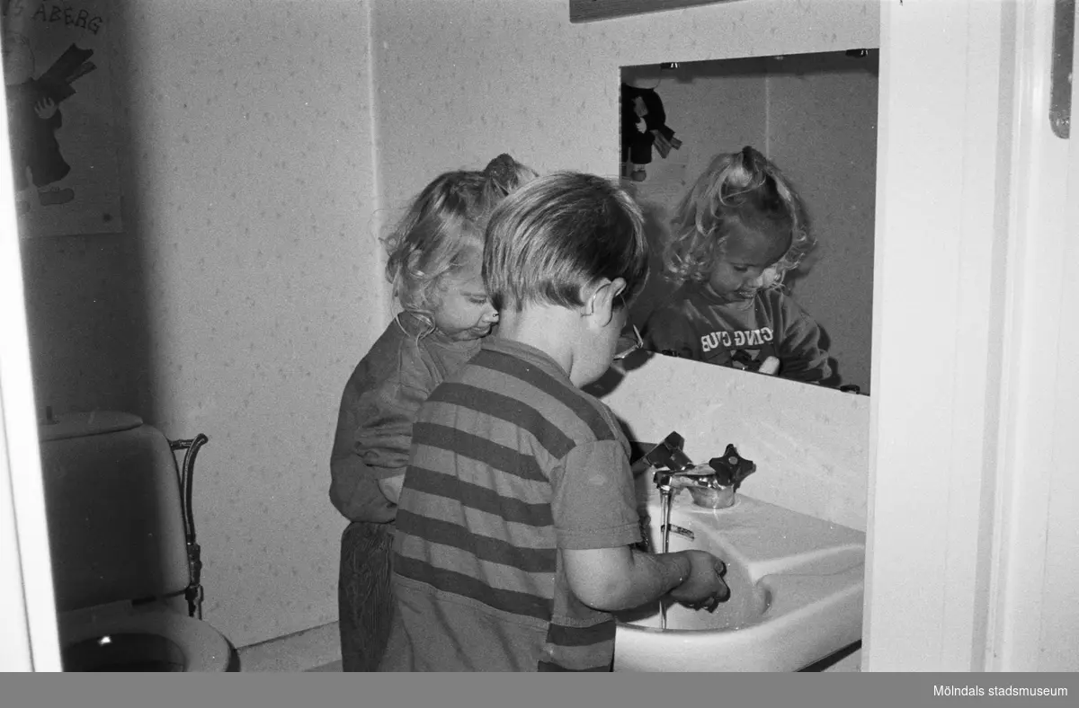 Jasmine och en pojke tvättar händerna i handfatet inne på en toalett. Barnen är fotograferade snett bakifrån, så man ser deras ansikten i spegeln, som hänger på väggen framför dem. Lille Skutt, Katrinebergs daghem, 1992.