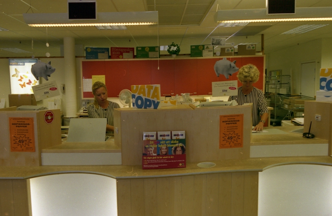 Två av kassorna och postkassörerna på regionpostkontoret Kungshörnet i Uppsala, 1996. På kassadisken finns ett broschyrställ.
