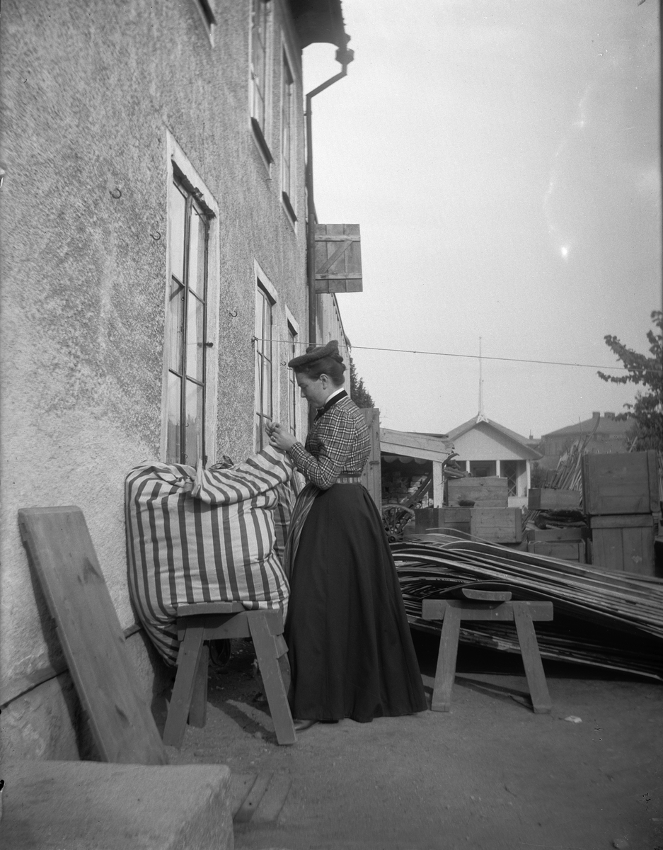 Elin Liljefors står utanför makens kruthandel och syr på ett bolster, Svartbäcksgatan 37, Svartbäcken, Uppsala 1901