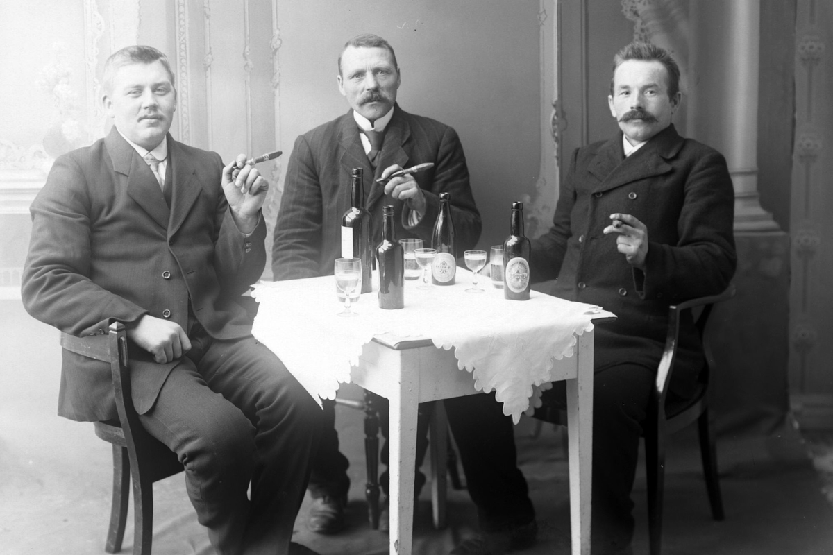 Studioportrett av tre menn som sitter rundt et bord med sigarer, øl og dram.