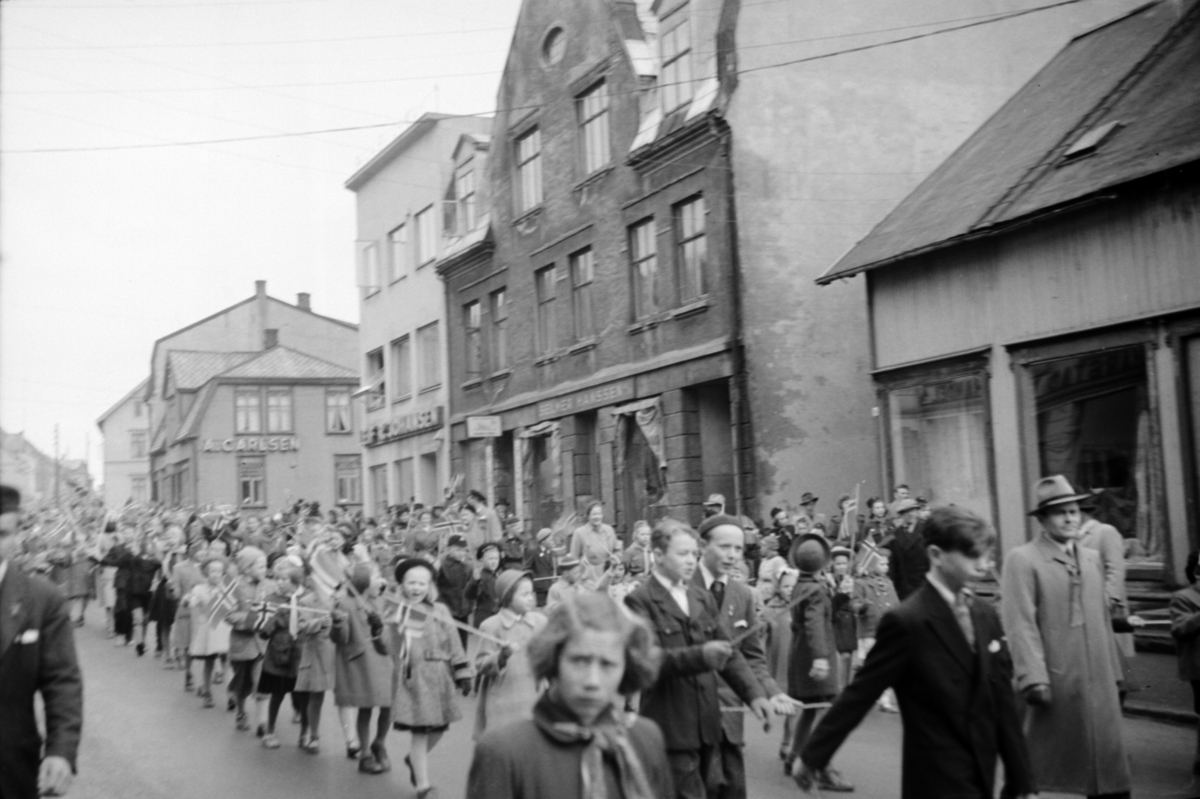 Barnetoget marsjerer ned Strandgata(?), på 17. mai. I bakgrunnen ser vi blant annet butikkene til A. Carlsen,  Leif Johansen og Selmer Hanssen.