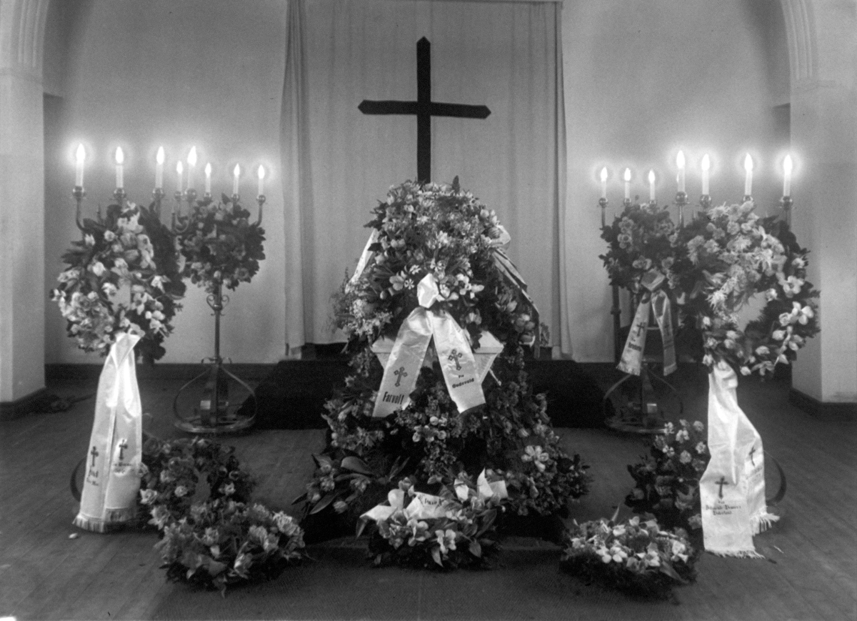 Båre i kirkerom pyntet med blomsterkranser og bånd. Fra originalprotokollen: "Simonsen. Gravferd".
