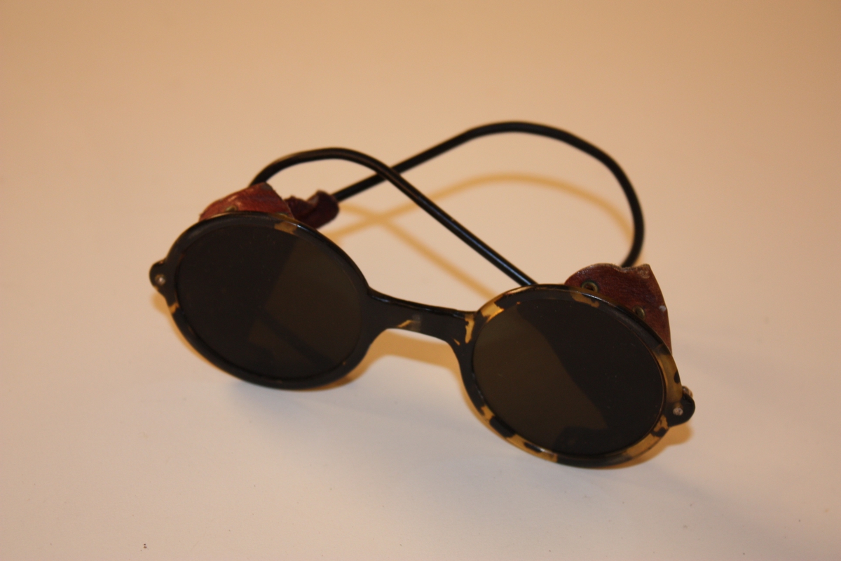 Rektangulært metallskrin. Runde briller med fleksible bogar. Skinnlappar på kvar side for å hindre solstråling frå sida.