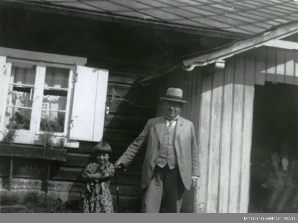 En liten jente med en katt i armene og en mann står ved en husvegg. Til venstre for dem er et vindu med skoddene åpne, til høyre et vindfang der vi kan skimte en person. Se også ST.K.HE 2007-011-0111 til -0126.