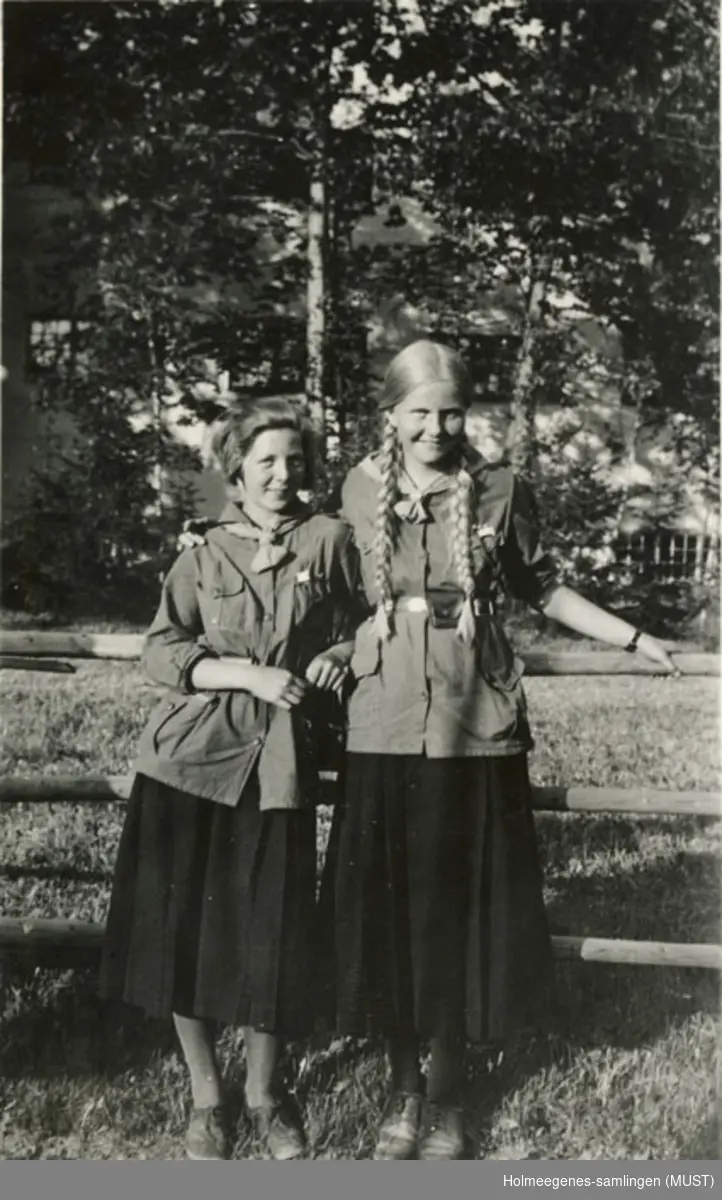 To speidere (jenter) i speiderdrakt oppstilt foran kamera. Et skogholt i bakgrunnen. Håndskrevet på baksiden av bildet: "Solveig Skavang og en venninde".