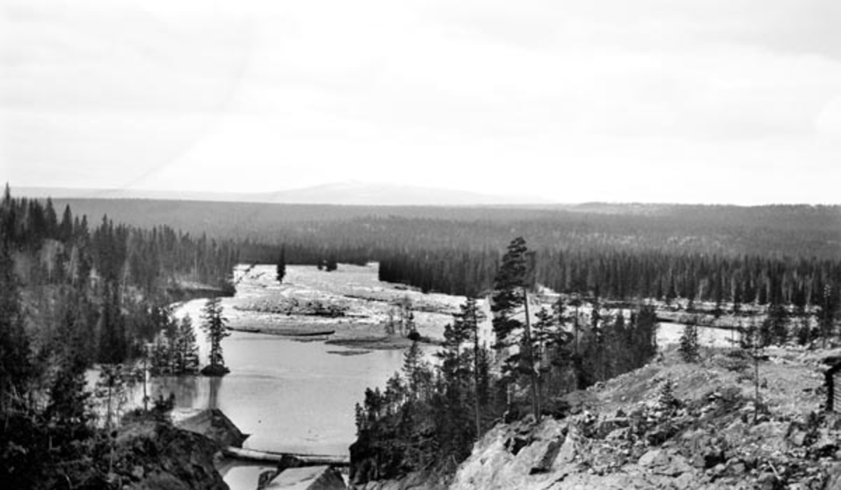 Utsikt fra Osfalldammen i elva Søndre Osa i Åmot kommune i Hedmark dagen etter det store dambruddet, som skjedde 10. mai 1916.  I forgrunnen ser vi ut over berget, der Søndre Osa opprinnelig passerte to avsatser med en samlet fallhøyde på 25,5 meter.  Det var dette fallet som i 1913-14 ble regulert med en dam på oversida og turbinrør til en nedenforliggende kraftstasjon med to turbiner som skulle levere strøm til belysning i sentrale deler av Åmot-bygda, samt energi til en ny treforedlingsbedrift («Rena Træsliperi», seinere «Rena Karton A/S»).  Våren 1916 - halvannet år etter at kraftstasjonen begynte å levere strøm - kom snøsmeltinga seint, men brått.  Med varmen kom dessuten vårregnet.  Følgelig ble kraftverksdammen raskt breddfull uten at de som skulle regulere dette vannmagasinet helt forstod å manøvrere lukene som skulle begrense belastningene på konstruksjonene.  Konsekvensen ble at vannet etter hvert begynte å renne over nordre damarm, der det grov seg et far som raskt ble større, slik at vannet fosset ut og rev med seg trær, sand, grus og steinblokker i det som raskt ble til et nytt elveløp nord for det opprinnelige.  Bildet viser hvordan terrenget så ut et par dager etter dambruddet, med et forholdsvis stillestående vannspeil sør for kraftstasjonen (som vi skimter deler av bak et par furutrær litt til høyre for sentrum av bildeflata).  Vi ser også det nye elvefaret lengre nord (som renner inn fra høyre på dette fotografiet), og vi ser hvordan den plutselige storflommen dambruddet utløste har revet med seg jordsmonn og skog i et bredt belte vestover mot Rena-elva.
