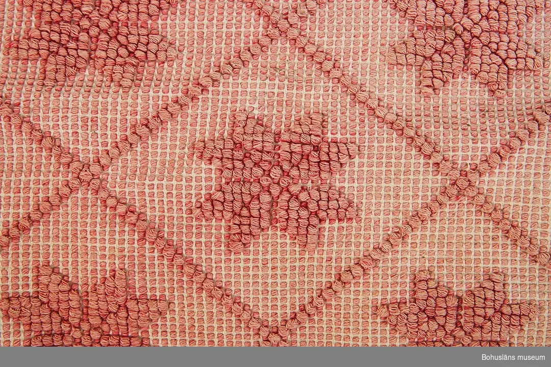 Avlångt täcke i noppväv i diagonalt rutmönster med en rund åttakantig blomma i varje ruta. Kantad runtom med rött bomullstyg. Ojämnt blekt.