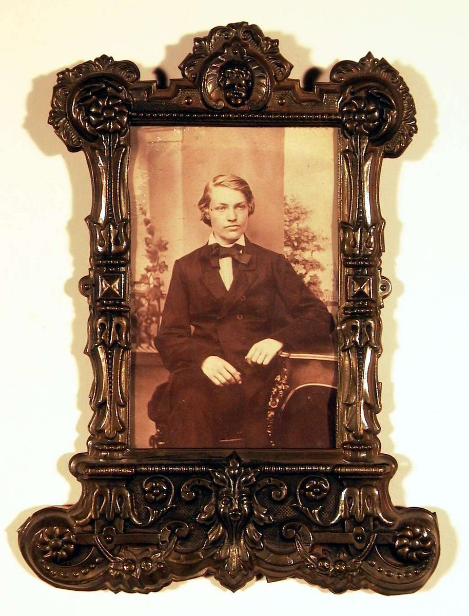 Portrettfotografi av ung mann med halvlangt lyst hår, sittende med benene i kors, kledd i mørk dress med halsbind.