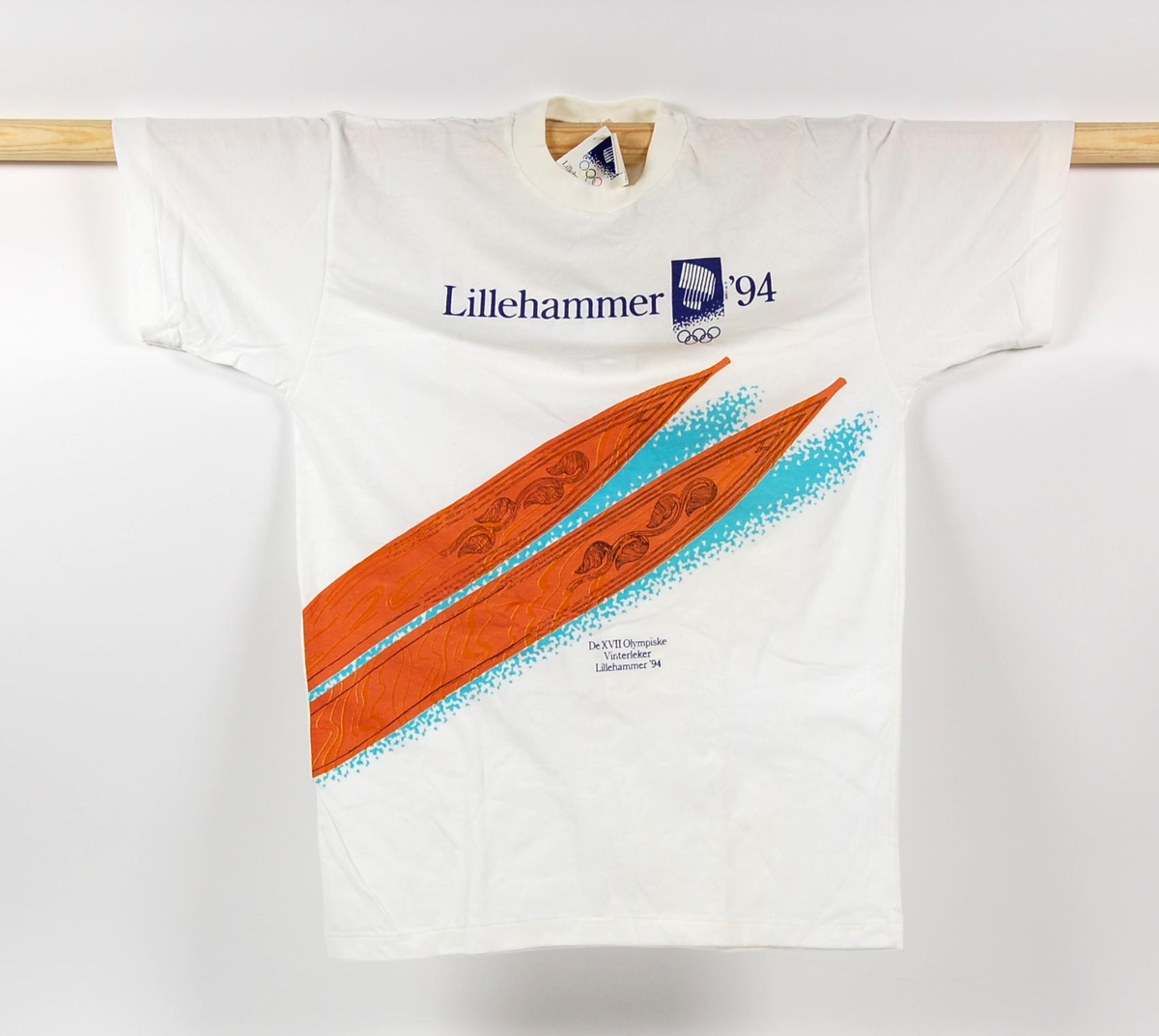 Hvit t-skjorte i størrelse M. Foran på t-skjorten er det en laminert logo for de olympiske vinterleker på Lillehammer i 1994 og et motiv av tuppene på et par treski av gammel modell.