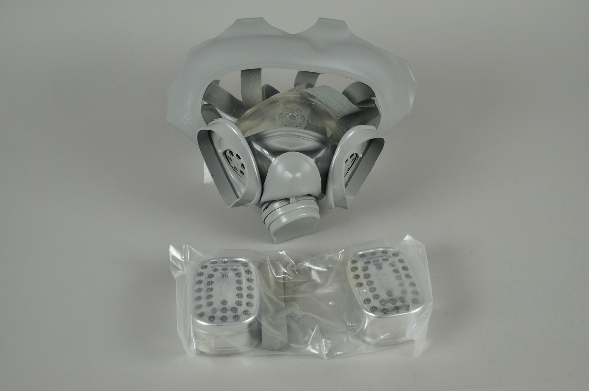 Gassmaske i plast med utskiftbare filter og regulerbare stropper for å feste over hodet. Veske til oppbevaring følger med.