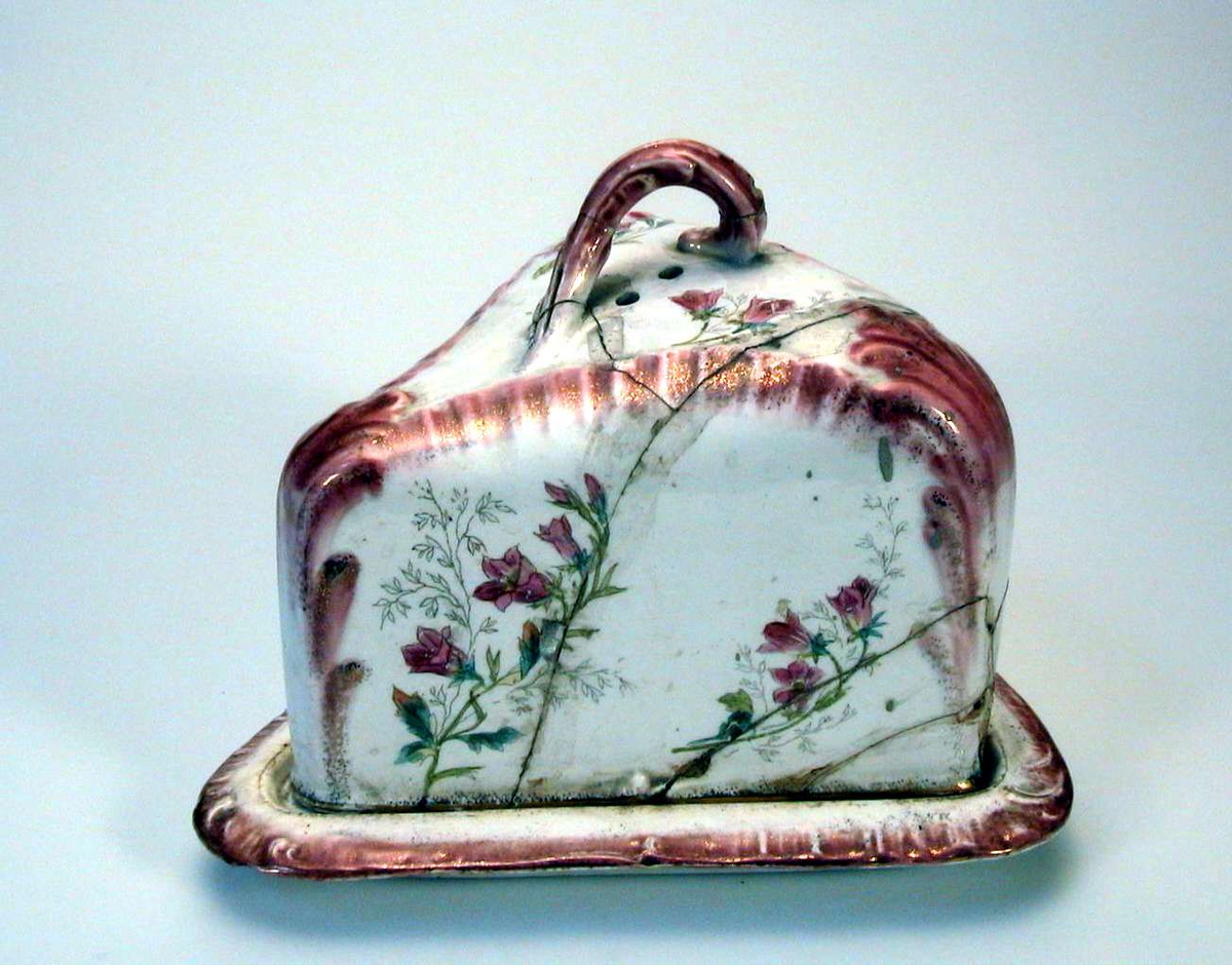 Trekantet osteklokke med skål og lokk. Den er i hvit keramikk med blomsterdekor i rosa og grønt, og med rester av forgylling. Håndkolorert dekor i trykket grunnmønster.