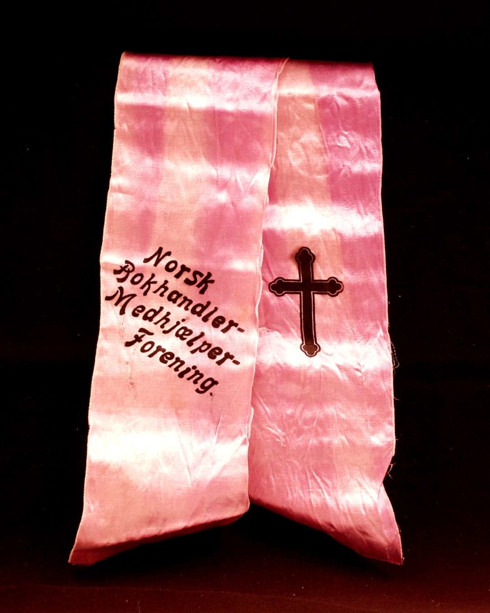 Bånd i lilla silke. Båndet er delt i to. På båndet står det: Norsk Bokhandlermedhjælperforening, sammen med et sort kors.