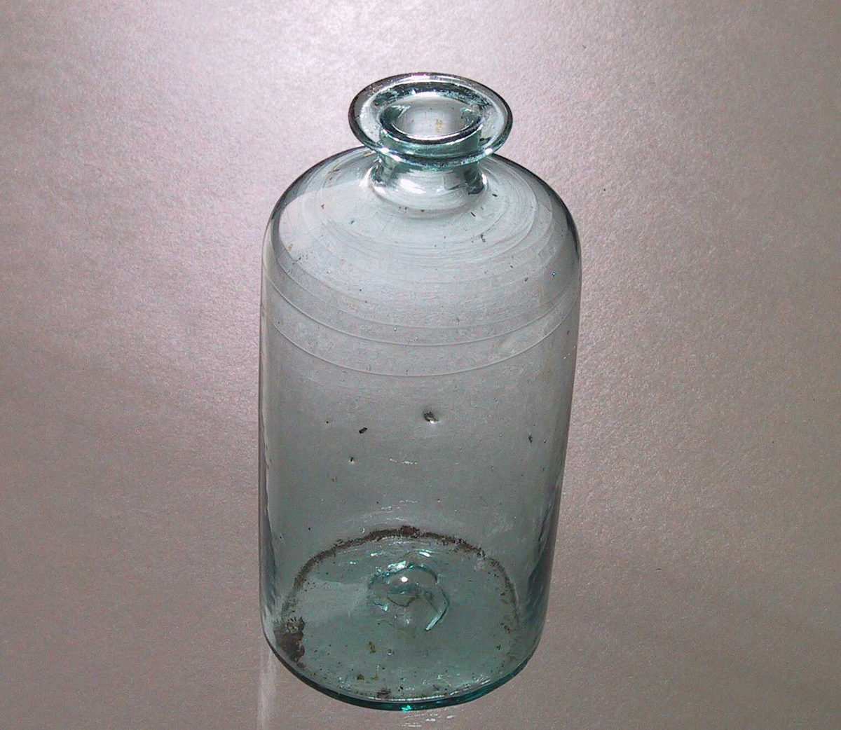 Flaske til medisin. Glass, grønnblått. Sylinderformet flaske med skrå skuldre, kort hals, utbrettet  munningsrand.  Kork mangler.