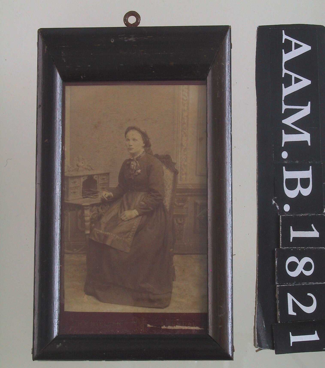Portrett av ung kvinne, Turid Homdrom, Gjøvdal, død 1936.. Sittende ved skrivebord.   Mørk kjole, forkle med mønstret bånd.  Mønstret sløyfe i halsen. Hodetørkle.  Bak interiør i nyrenessanse.  