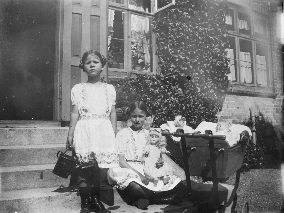 To jenter på trappen foran et hus, med dukker og dukkevogn.