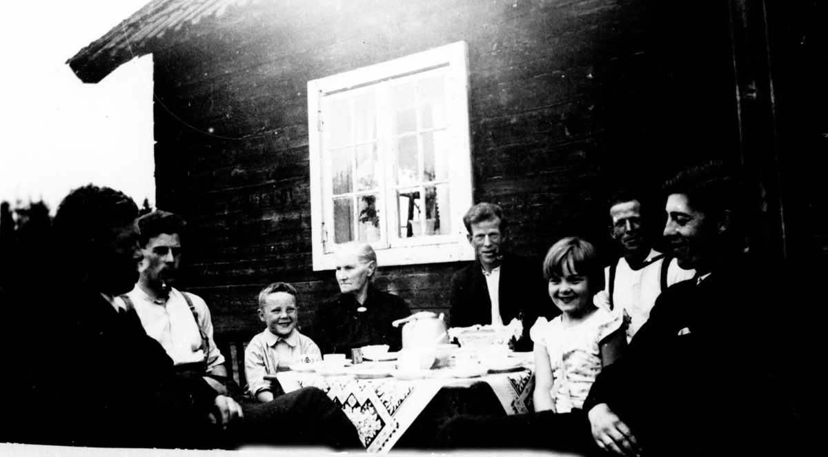 Husmannsplass. 5 menn, 3 røyker pipe, 1 eldre kvinne, en gutt og en pike samlet rundt dekket kaffebord utenfor huset.
