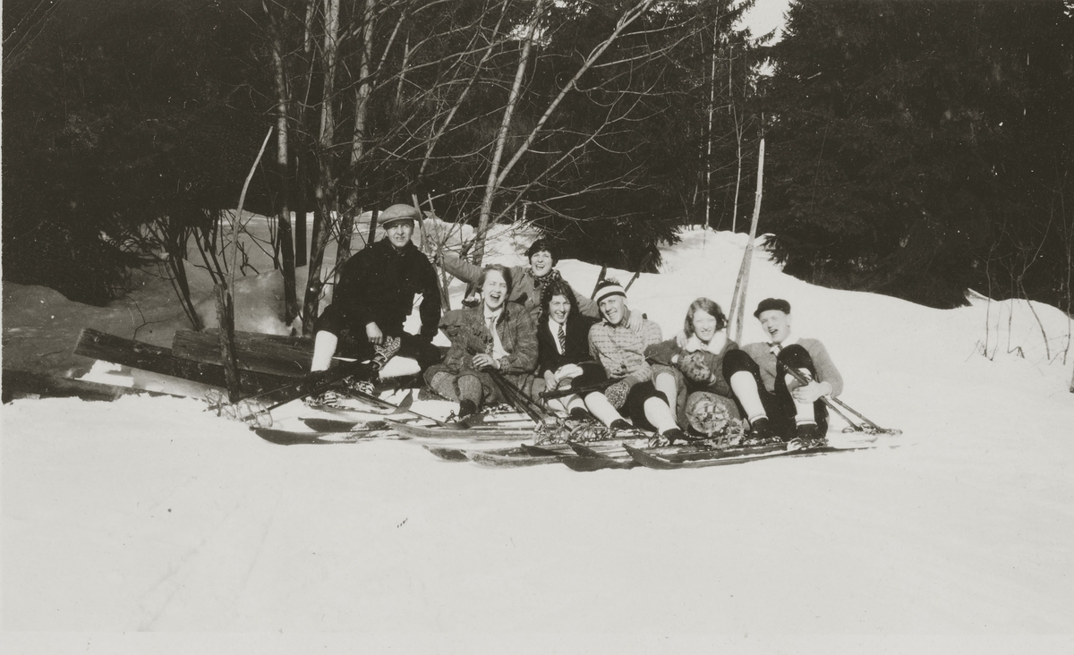 Fire damer og tre menn sitter i solen med ski på bena.