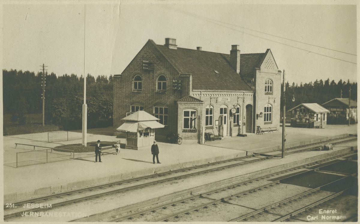 Jessheim jernbanestasjon. Stasjonsmesteren og reisende står på plattformen.