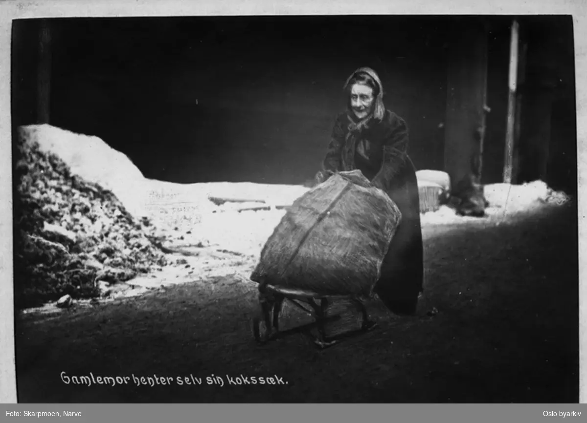 En gammel kvinne frakter en kokssekk på en kjelke (liten spark?), vinter med snø i gata.
