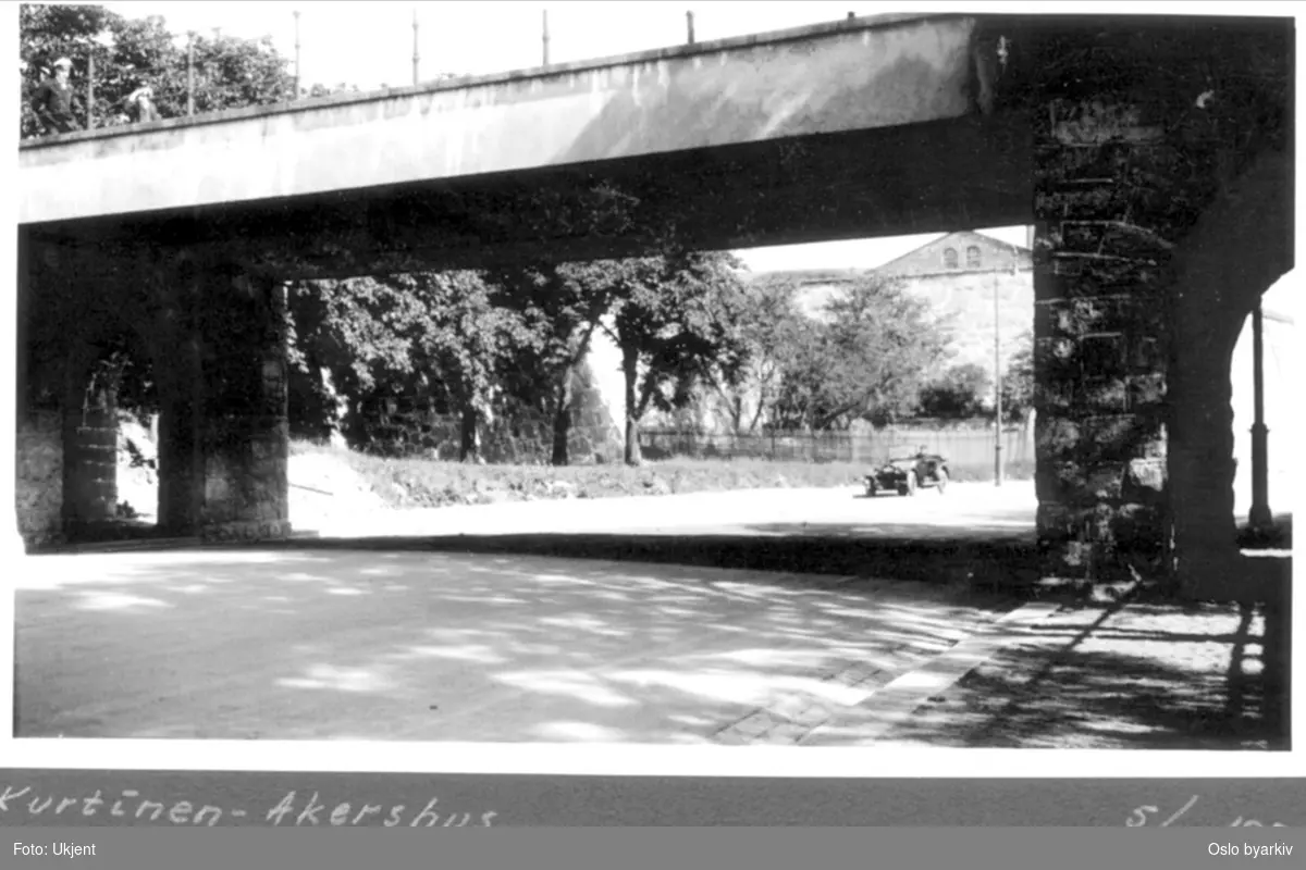 Kongens gate nordover med festningsbrua (Vindebrua) og to uniformerte menn som passerer. Trær langs vollen.Trestakitt i hjørnet (Frukttrær) under Halmlageret . 1920-talls bil nedover gata. Bildet tatt 5 Juli.
