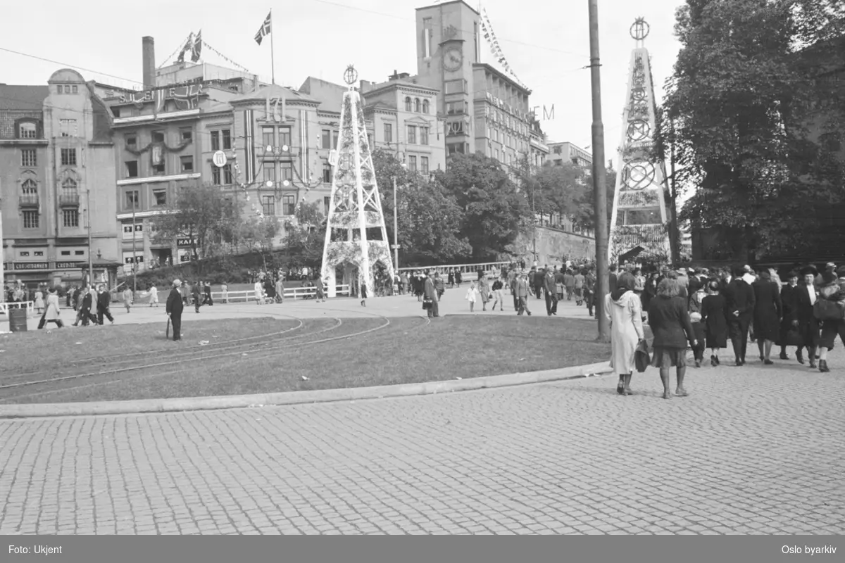 Østre Rådhusplassen. Folk på vandring på tidligere Tordenskiolds plass og Rådhusgata. Utsmykkede piramidetårn og pyntede bygninger i anledning Kongens hjemkomst 7. juni 1945.