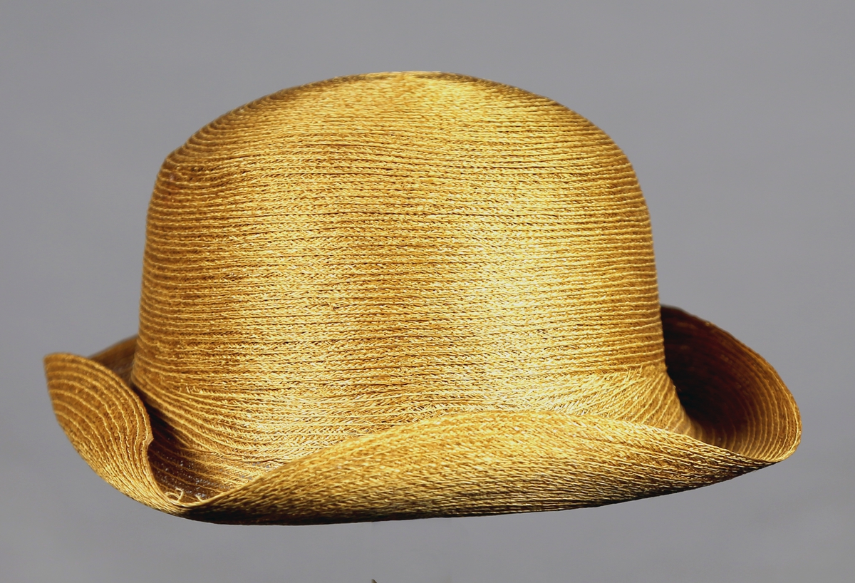 Hatt ("presset capelin") med rund og stiv pull, og brem som er brettet opp. Hatten er av sammensydde stråbånd