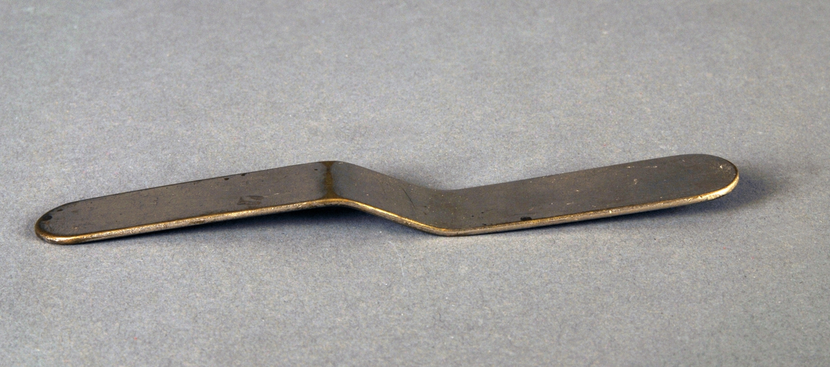 Et tynt, smalt og flatt metallstykke, endene er avrundet. En knekk midt på. Ene halvdel er noe bredere enn den andre.