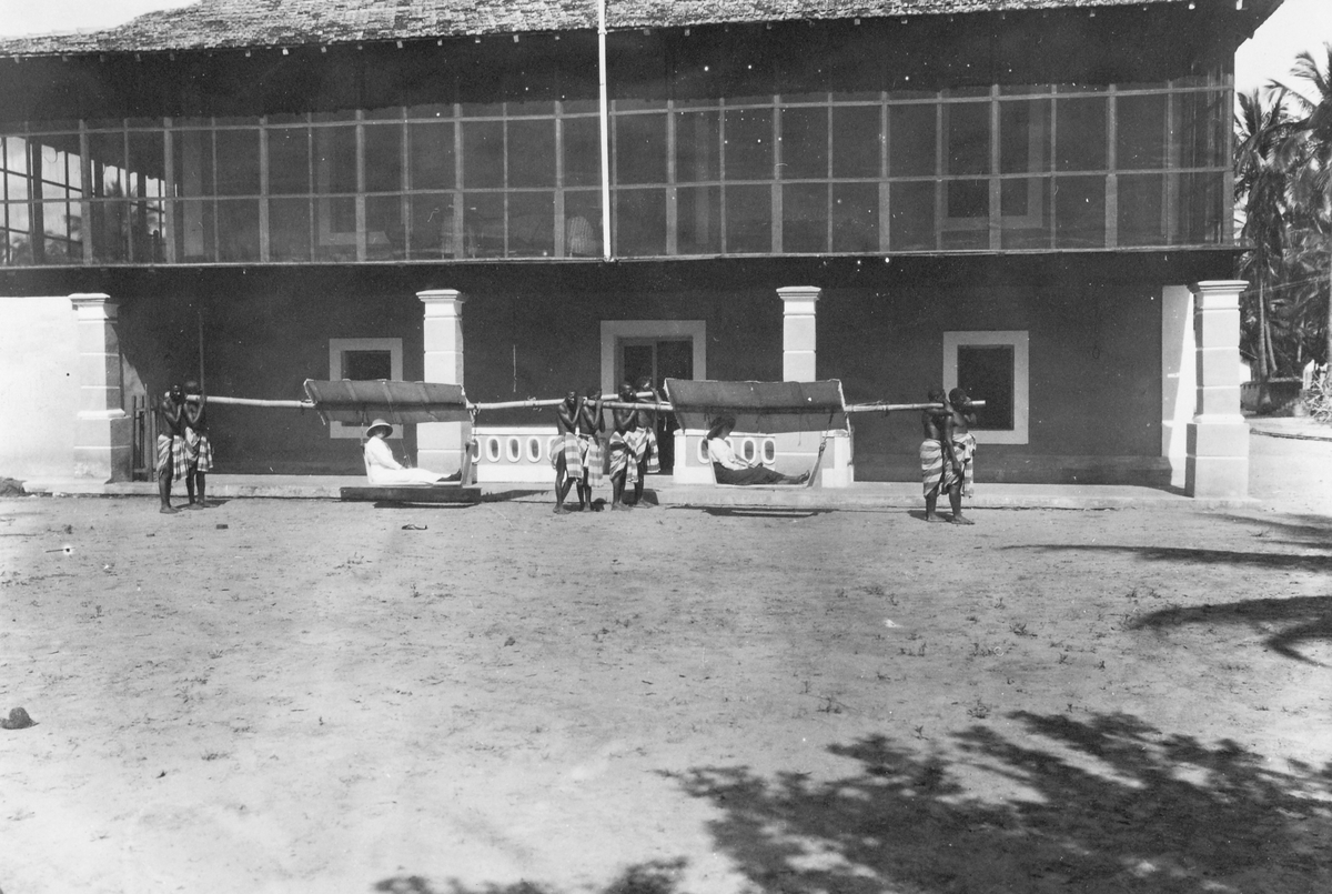 Mosambik. 1914. To damer i bærestoler foran en større bygning.