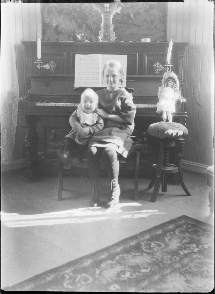 Interiør, stue. Jente med en baby på fanget sitter foran et piano og en dukke. 