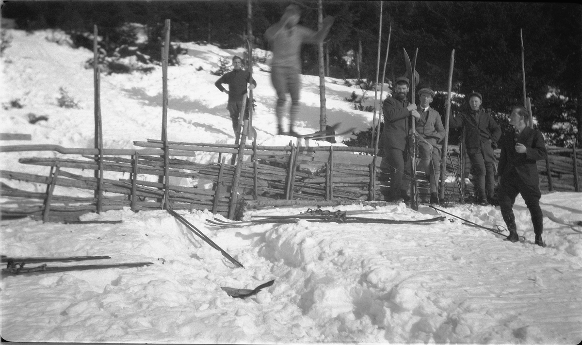 Ukjent mann hopper på ski over en skigard. Gruppe menn ser på. 