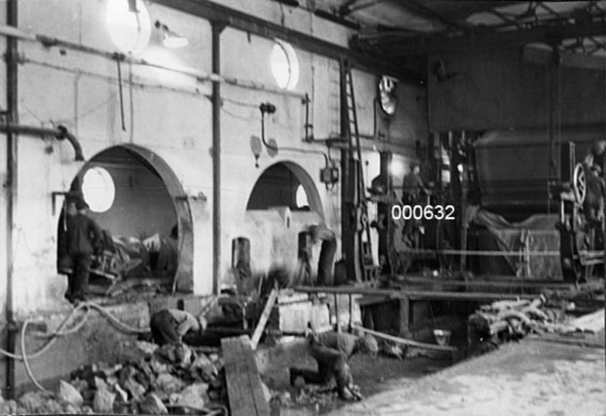 Interiør fra maskinsalen ved A/S Buskerud Papirfabrikk på Åssida i Drammen (den gang i Lier kommune).  Fotografiet er tatt under ombygging av papirmaskinen i 1937. Det viser en sal der maskinens våtparti er fjernet.  Også deler av det støpte golvet er fjernet, antakelig med sikte på å muliggjøre videre nedgraving for å oppnå større dybde for en ny maskin.  Ei lita steinrøys fra dette arbeidet ligger ennå på marka der hvor våtpartiet har stått.  Her står to arbeidere bøyd over et arbeid.  Til venstre ser vi veggen mot maskinrommet, med diverse rundbuete åpninger for akslinger m. m.  Høyere opp på veggen er det sirkelrunde vinduer som slapp dagslys inn i rommet.  Tilsvarende runde vinduer skimtes også i maskinrommet, gjennom de rundbuete åpningene i vegge.  I bakgrunnen til høyre er enden av papirmaskinens tørkeparti tildekt med presenninger.  