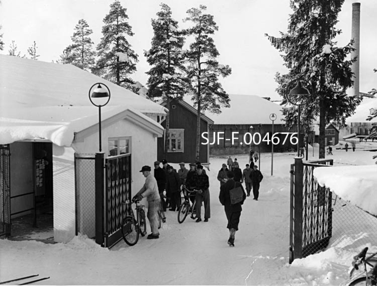Fra porten til Drammenselvens Papirfabrikker på Modum i Buskerud.  Fotografiet er tatt ved arbeidstidas slutt en marsdag i 1957.  Vi ser en del arbeidere på veg ut, noen med sykler, andre gående.  Fabrikkområdet var åpenbart omgitt av et høyt nettinggjerde, og porten var en svartlakkert, todelt jernkonstruksjon med fiskebeinmønstret rammeverk.  Ved siden av porten ligger den kvitmalte murbygningen fra 1954, som inneholdt bad og vaskeri for de ansatte og deres familier, samt som kontor for portvakta.  Vi ser også at det var ei spesiell «sluse» foran portvaktrommet, der det hang et stemplingsur som mange av de ansatte måtte bortom.  Bak og til høyre for den kvite murbygningen ser vi gavlen på en panelt trebygning som lenge ble brukt som spisebrakke for fabrikkens ansatte, seinere som elektrokontor.  Bak denne igjen sto den store papirlagerbygningen med skyveporter mot jernbanesporet, som gikk langs langveggen mot innkjøringsvegen til fabrikken og elva.  Lengst til høyre i bildet skimter vi en av de teglstensmurte maskinsalene og den høye, sylindriske fabrikkpipa. 