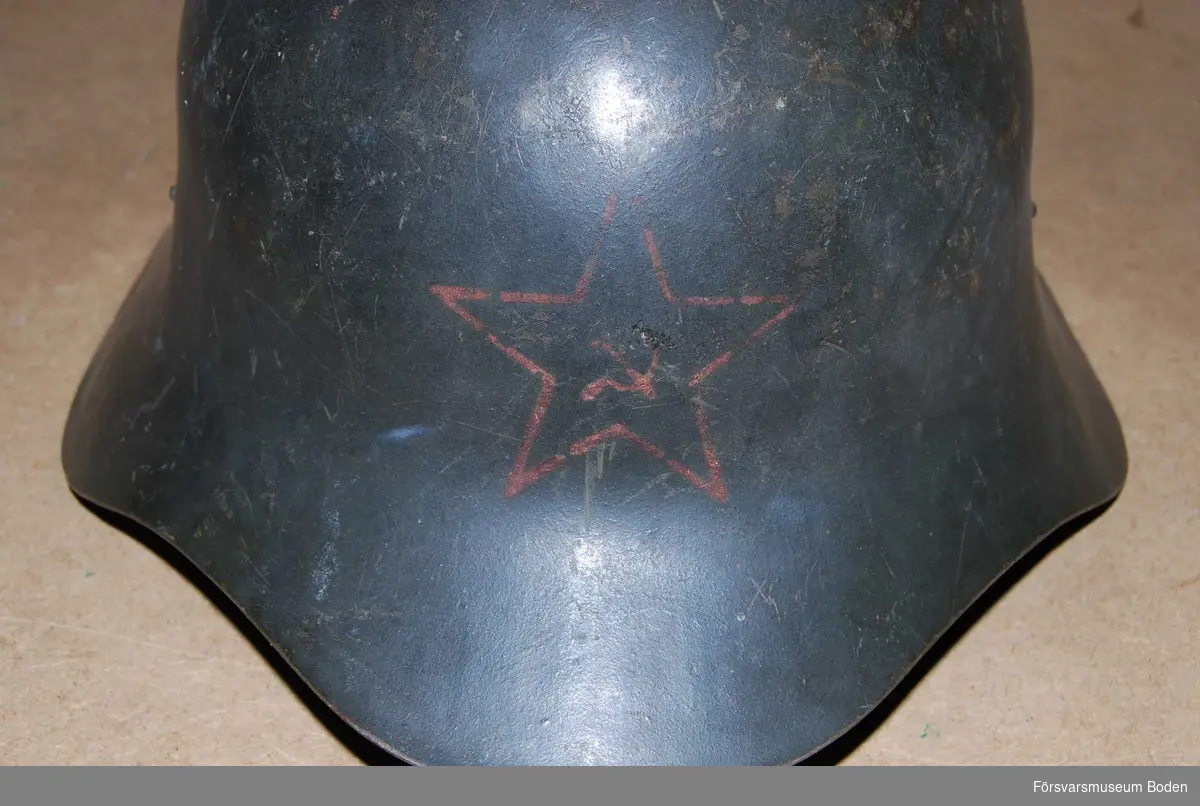 Svartgrön stålhjäm med hakband men utan inredning. Framtill finns en röd stjärna med hammare och skära i mitten.