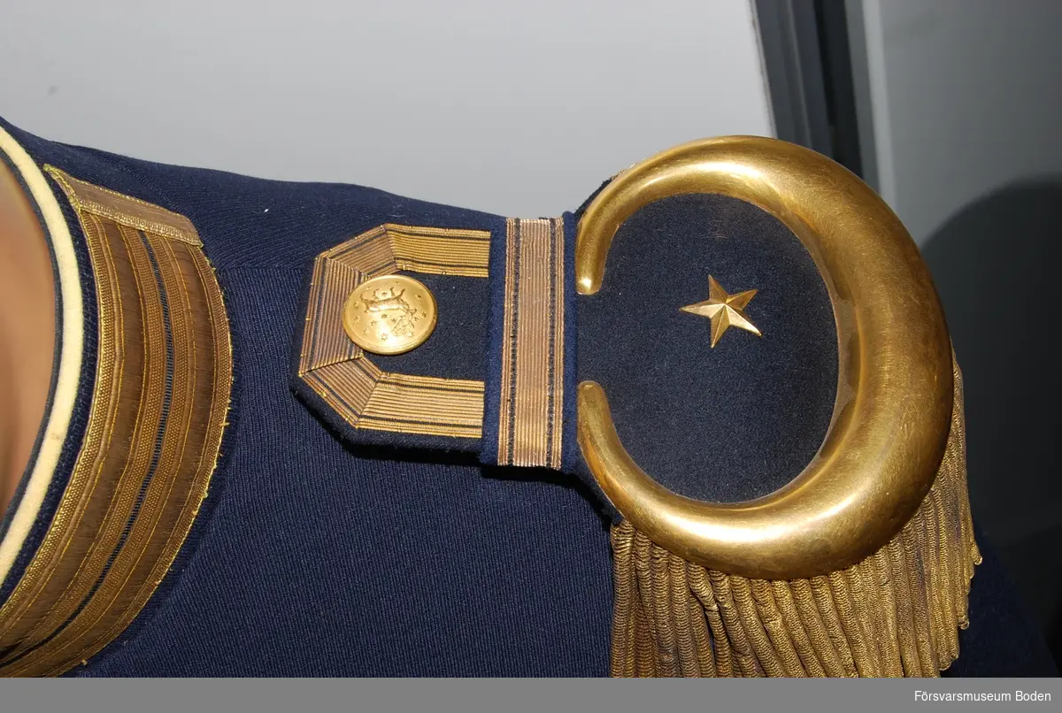 Av modell I som användes vid infanteriregementena. Mörkblått kläde med krage i samma färg, försedd med dubbla knapphålsgaloner i guld. Gult foder och gula passpoaler runt krage och kanten framtill (nu kraftigt blekta). Knäppt med 8 knappar av regementets m/1893 i guld. Utrustad för paraddräkt med epåletter, fastsatta med slejfar försedda med guldgalon. Epåletterna har en metallstjärna tillsammans med buljoner (fransar) vilket betecknar major. Ärmuppslag med dubbla stående knapphålsgaloner i guld samt regementets knappar. Gula passpoaler längs skörtets kanter baktill. Skörtet försett med tre knappar i dubbla rader. Buren av Olof Dahlqvist.