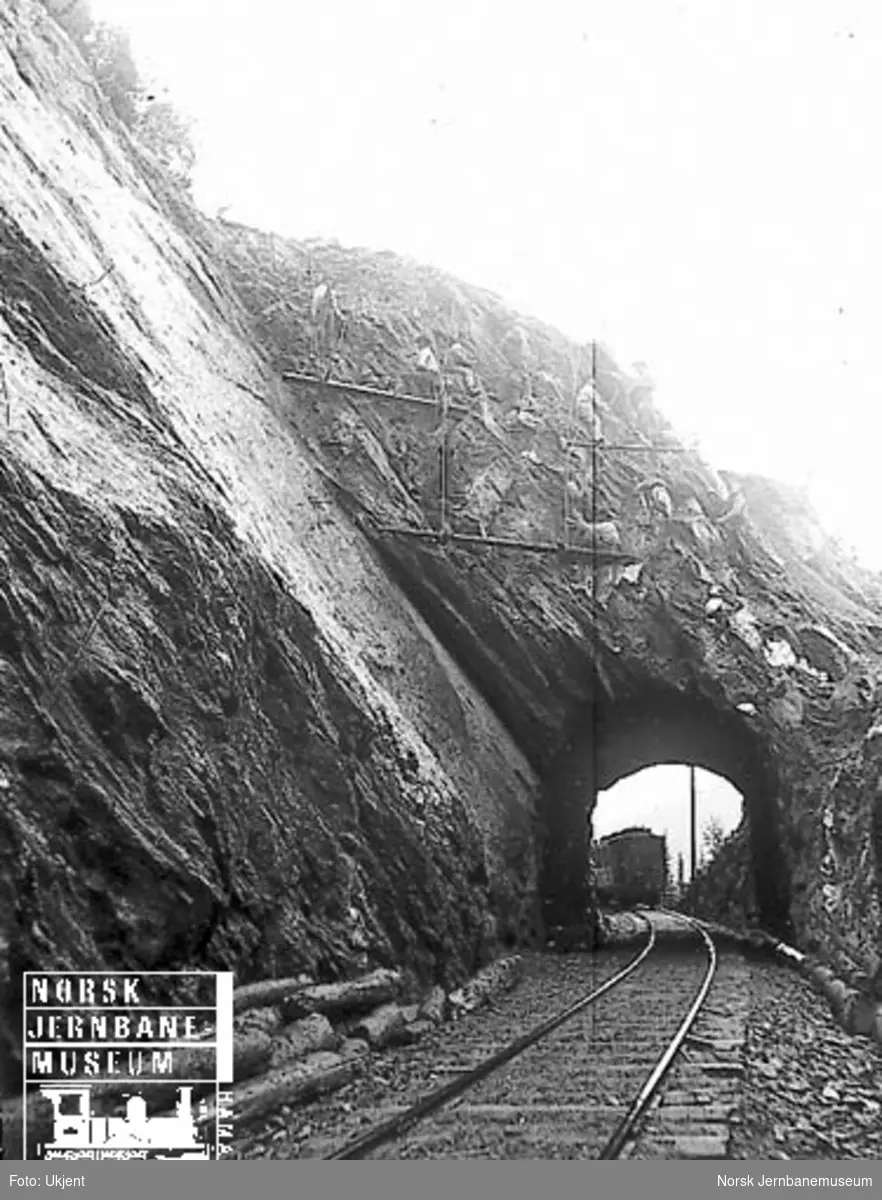 Ni menn utfører fjellsikring ved en kort tunnel, trolig på Flekkefjordbanen : personvogn på motsatt side av tunnelen