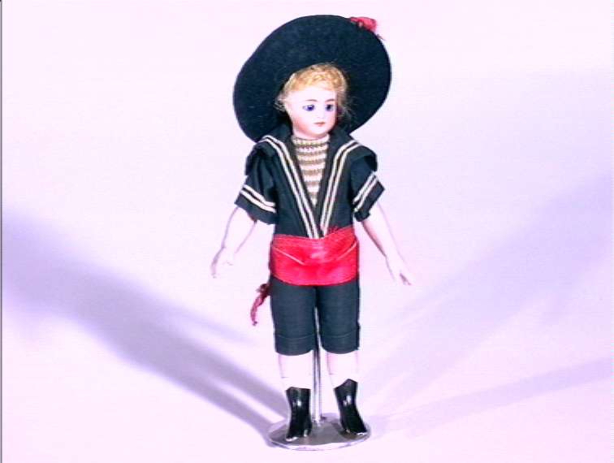 Dukkegutt med matrosklær, bredbremmet hatt og rødt magebånd. Hode, armer og ben i porselen.