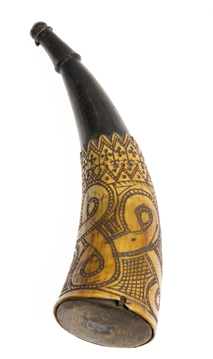 Krutthorn med flat bunnplate i tre. Ornamentikk på horn i form av to horisontale bårder hvorav den tykkere er fylt med et bånd som går i løkker rundt hornet mot en skravert bakgrunn. Den andre bården er fylt med grafiske mønster.