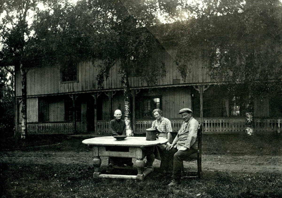 Hofsrud, Øvre Romerike, Nes, Akershus. To kvinner og en mann sitter ved bord på gårdsplassen foran våningshus med svalgang.
