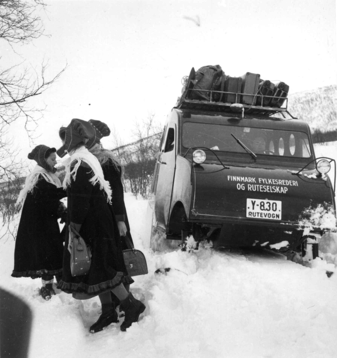 Snowmobile. Snøbil med oppakking på taket sitter fast i snøen,ved siden av står tre samekvinner. Karasjok 1958. Denne beltevogna var registrert Y-830 med undertekst "Rutevogn". Den gikk i rutetrafikk hos Finnmark Fylkesrederi og Ruteselskap (FFR).