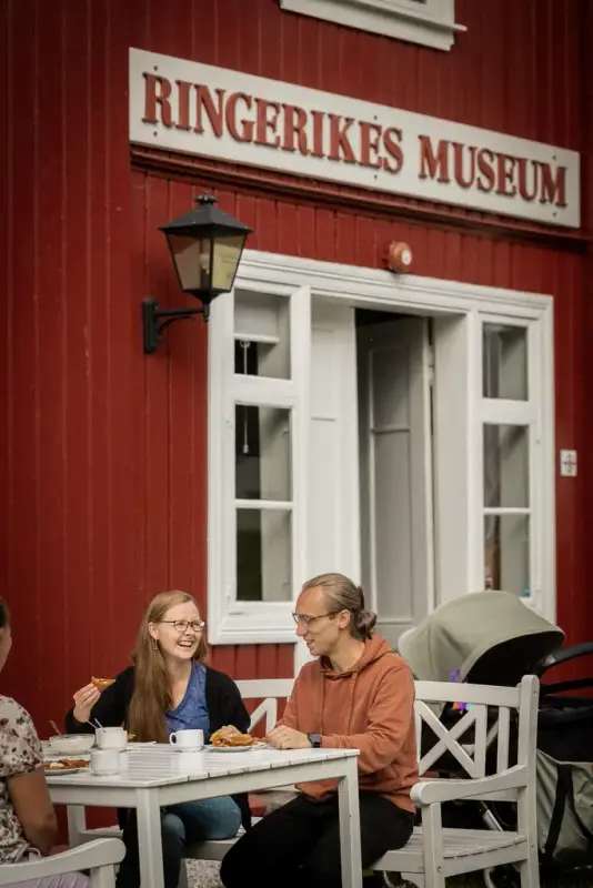 Bildet viser fasaden til Ringerikes Museum, med en rød bygning og hvite detaljer. Utemøbler er plassert foran inngangen, og to mennesker sitter og hygger seg med vaffel og kaffe. Bak dem er en barnevogn. .