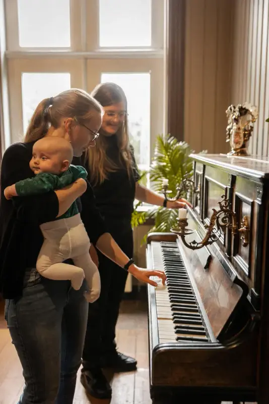 En ung kvinne med en baby på armen viser entusiastisk frem hvordan man spiller på et gammelt historisk piano. I bakgrunnen står en annen kvinne og følger smilende med. 