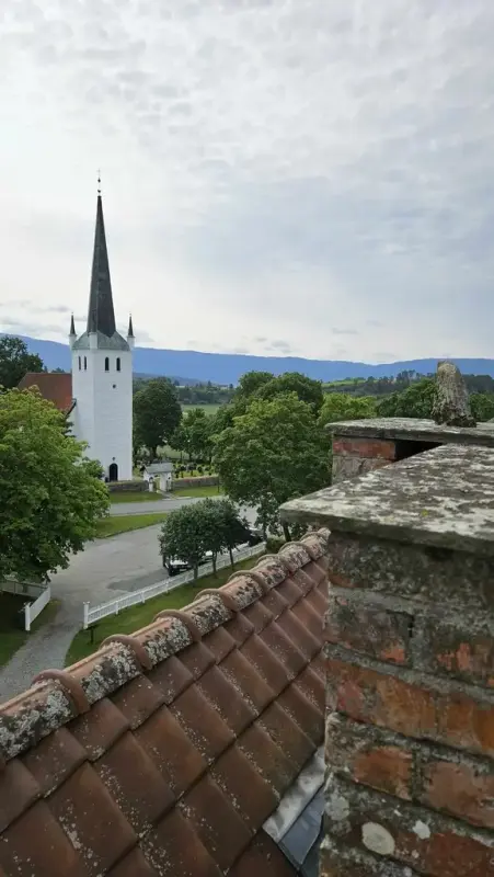 Bildet viser Norderhov kirke - en hvitkalket middelalderkirke i stein med høyt mørkegrønt spir. Bilde er tatt fra taket på Ringerikes Museum og i bildets forgrunn er det rød takstein med mose. 