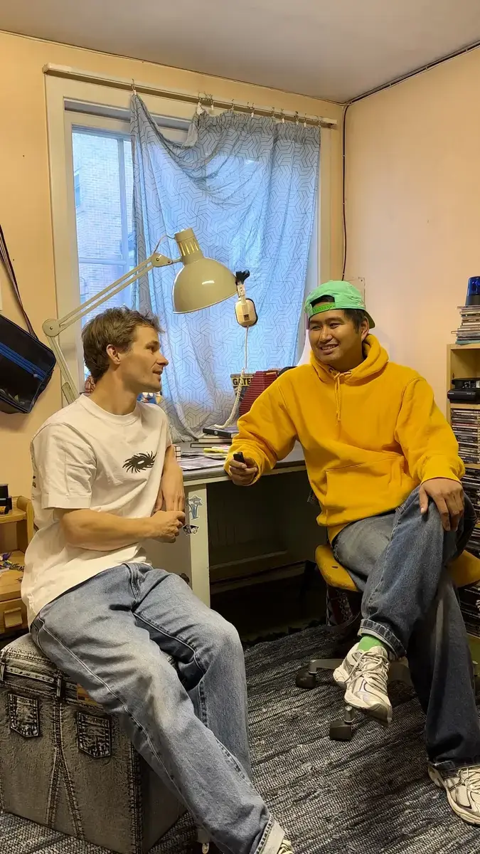 To menn sitter og snakker sammen i et rom som fremstår litt rotete. De er kledd i avslappede hiphop-klær